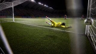 Action rear view of a female soccer goalie diving and narrowly missing the ball before it goes in the goal