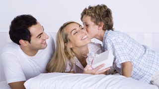 4020d6c5-Woman lying in bed with husband and son, holding gift, boy kissing her cheek