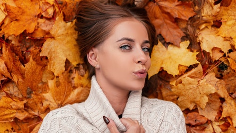 Happy chic pretty young woman with blue eyes with brown hair in a knitted fashionable sweater is resting lying on the orange leaves in the park. Stylish girl with a beautiful smile. View from above