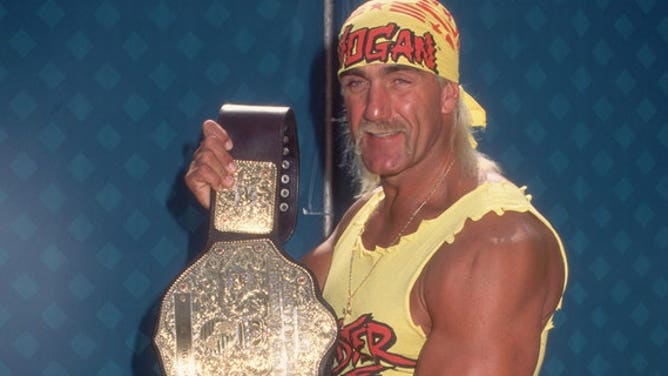 Hulk Hogan displaying his championship belt. Hogan wears a costume consisting of a yellow 'HOGAN' bandana, a yellow muscle top, a weightlifter's belt, red spandex pants, and yellow cowboy boots. 