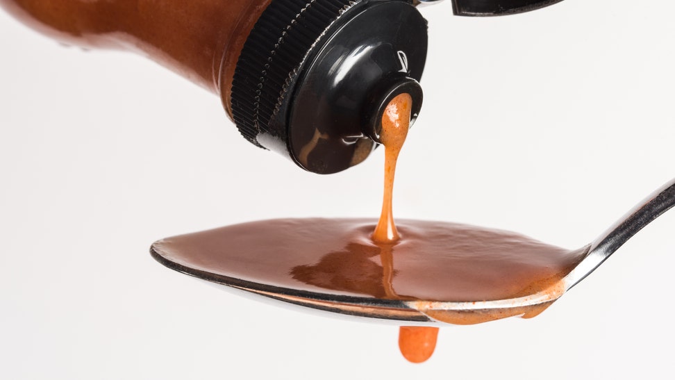 Close-Up Of Bottle Pouring Hot Sauce In Spoon Against White Background