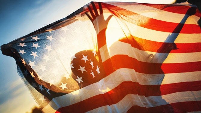Closeup rear view of a couple holding American flag against summer sunset. They are visible as silhouettes.